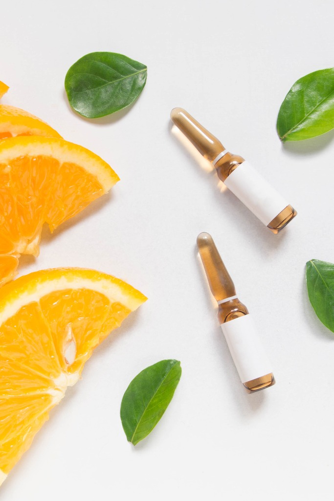 Draufsicht auf eine braune Vitamin C-Ampulle zur Injektion mit frischen saftigen Orangenfrüchten und grünen Blättern auf einem weißen Tisch. Vitamin synthetische oder natürliche Ernährung Entscheidungen Konzept. Attrappe eines Schönheitsprodukts.